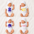Four newborn babies lying on a white textured blanket, each wearing a white bodysuit and hat with a different graphic design: a purple octopus with an exclamation mark, a yellow crescent moon with the words 'Snuggle Time', a purple elephant with a hanging cord, and the word 'Hello' in pink script.