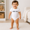 A baby standing on a white rug in a bright nursery, wearing a white short-sleeve bodysuit with 'BABY LOVE' printed in black across the chest.