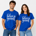 A man and a woman standing side by side wearing matching royal blue t-shirts with the phrase 'believe in yourself' printed in white script across the chest, paired with blue jeans against a light gray studio background.