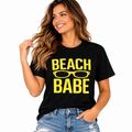 A woman wearing a black short-sleeve graphic t-shirt with 'BEACH BABE' and sunglasses printed in yellow, paired with distressed blue jeans, against a solid white background.