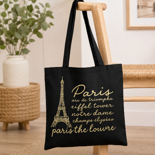A black canvas tote bag with gold printed text and an Eiffel Tower graphic, hanging on a wooden chair in a softly styled indoor setting.