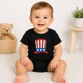 A smiling baby sitting on a white carpet wearing a black short-sleeve bodysuit with a red, white, and blue patriotic top hat graphic on the chest. A teddy bear is in a basket on the left, and a small plant on a wooden stool is on the right.