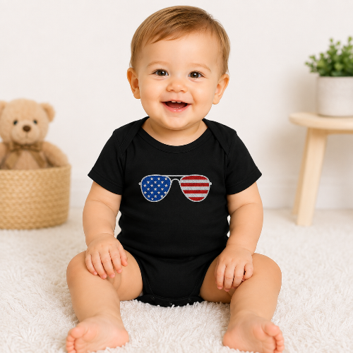 A smiling baby sitting on a white fluffy rug wearing a black short-sleeve bodysuit with a graphic of sunglasses featuring an American flag pattern. The background shows a cozy nursery setting with a teddy bear in a basket and a small wooden stool.