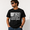 A man wearing a black crewneck t-shirt with a white distressed American flag graphic and a jumping bass fish design, set against a light textured wall.