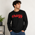 A man wearing a black long-sleeve crewneck sweatshirt with the word 'crazy' printed across the chest in red cursive script, paired with blue jeans, standing against a light gray wall.