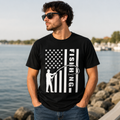 A man wearing a black t-shirt with a white graphic of a fishing flag and the word 'FISHING' stands in front of a rocky waterfront.