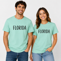 A man and a woman standing side by side wearing matching mint green t-shirts with the word 'FLORIDA' printed in black across the chest, paired with blue jeans, against a plain white studio background.