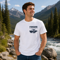 A man wearing a white crewneck t-shirt with a dark navy graphic that reads 'FOCUS IN YOUR MOUNTAINS' above a mountain and pine tree illustration, standing outdoors in front of a scenic mountain river landscape.