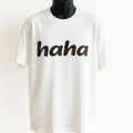 White graphic t-shirt featuring the word "haha" in bold black lettering, funny minimalist unisex graphic tee for casual everyday wear.
