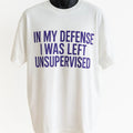 White graphic t-shirt featuring the humorous phrase "In My Defense I Was Left Unsupervised" in bold blue typography, funny unisex graphic tee.
