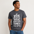 A man wearing a dark gray graphic t-shirt with the white printed text 'if you are going THROUGH difficult TIMES keep going', paired with blue jeans, against a plain light gray background.