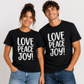 A man and a woman standing side by side, both wearing matching black crewneck t-shirts with the words 'LOVE PEACE JOY!' printed in large white block letters across the front, paired with blue jeans against a light gray studio background.