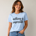 A woman wearing a light blue graphic t-shirt with the phrase 'nothing is impossible' printed in black script across the chest, paired with blue jeans, set against a plain light gray background.