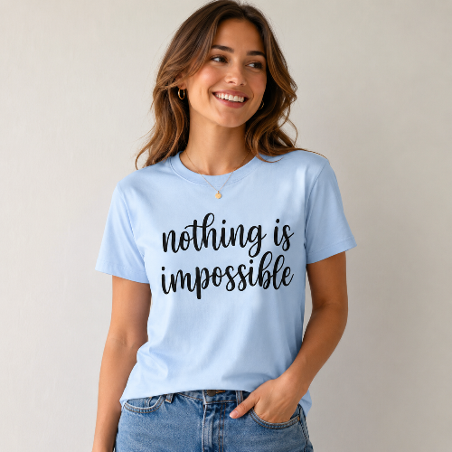 A woman wearing a light blue graphic t-shirt with the phrase 'nothing is impossible' printed in black script across the chest, paired with blue jeans, set against a plain light gray background.