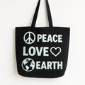 Black cotton tote bag with Peace Love Earth white graphic design featuring peace sign heart and globe symbols, reusable shoulder tote for shopping, travel, school, eco-friendly lifestyle, everyday carry, and sustainable fashion accessories