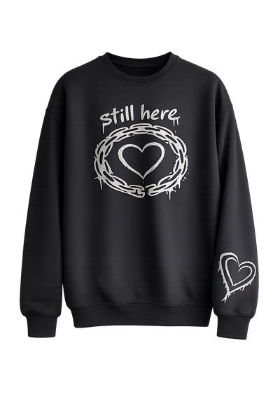 Black unisex crewneck sweatshirt featuring the phrase "Still Here" with a heart graphic on the front and a matching heart detail on the sleeve. Soft fleece everyday sweatshirt.