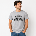 Heather gray unisex short sleeve t-shirt featuring a black Wild Horizons mountain adventure graphic with evergreen trees, sunrise, and outdoor typography on the front.