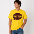 A man wearing a bright yellow t-shirt with a dark brown speech bubble graphic on the chest that reads 'WHAT'S UP?' in yellow letters, paired with blue jeans, against a plain light gray background.
