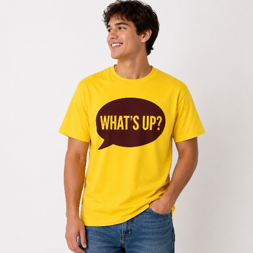 A man wearing a bright yellow t-shirt with a dark brown speech bubble graphic on the chest that reads 'WHAT'S UP?' in yellow letters, paired with blue jeans, against a plain light gray background.